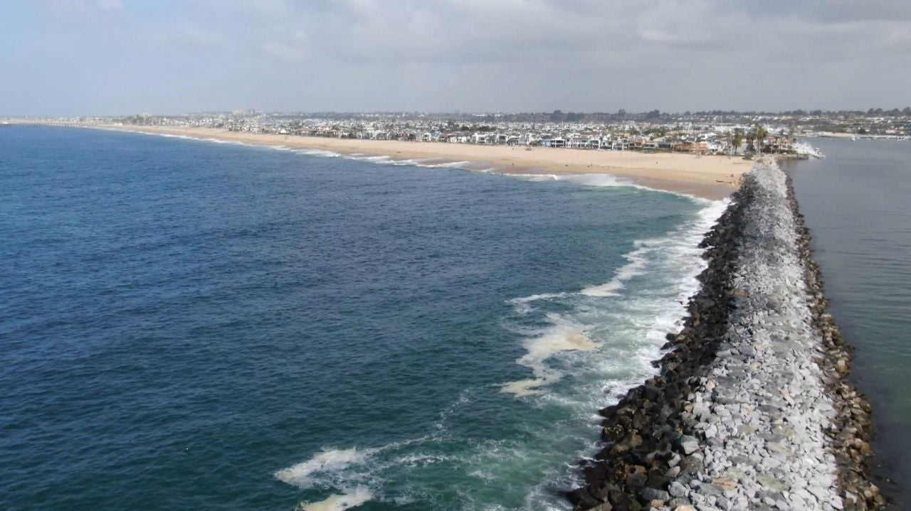 Aerial view of Newport Beach jetty with ocean waves, sandy shore, and coastal homes.