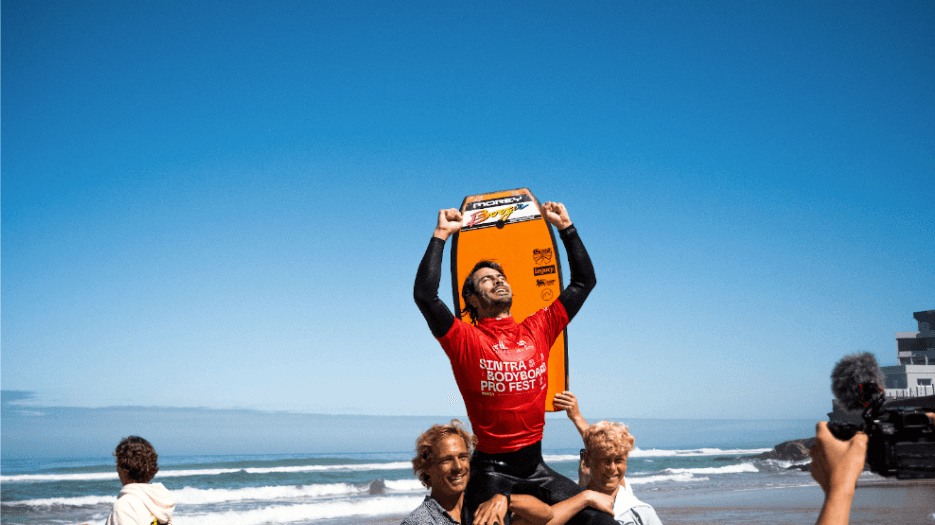 Bodyboarder celebrating victory on beach, lifted by friends, holding orange bodyboard