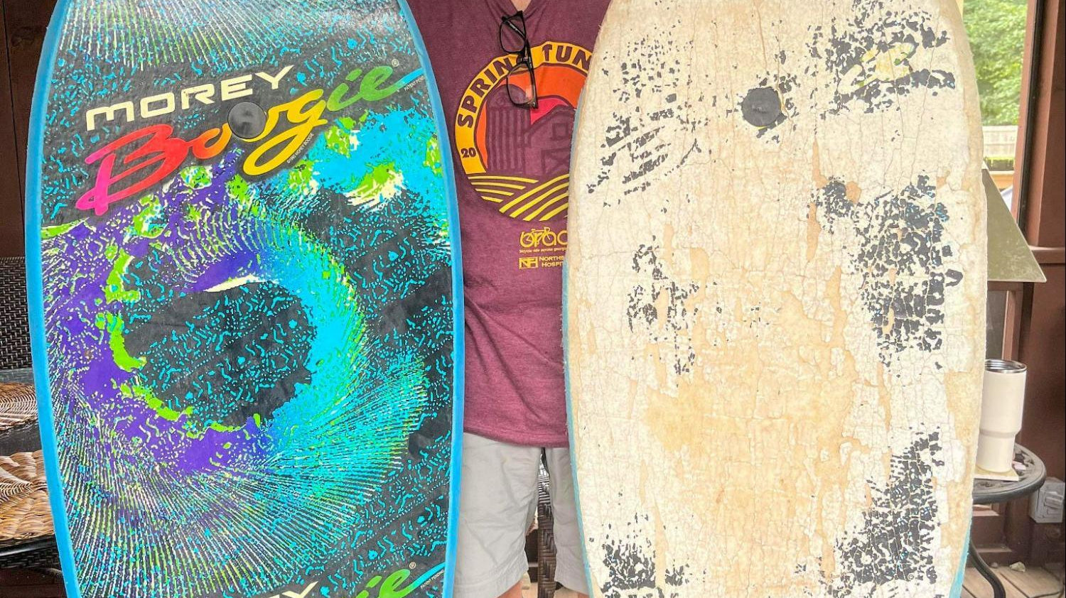 Man indoors holding two bodyboards, one new colorful Morey Boogie and one worn, with a black dog nearby