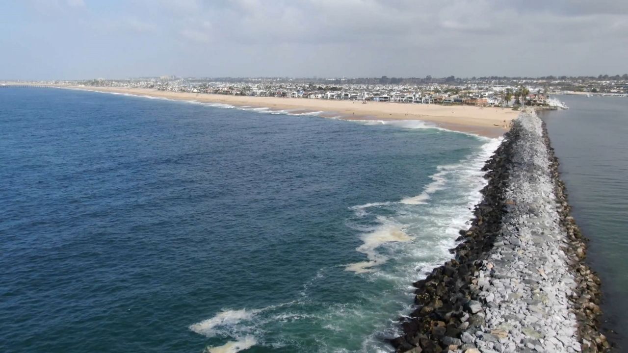 Aerial view of a rocky jetty extending into the ocean with a sandy beach and coastal city in the background