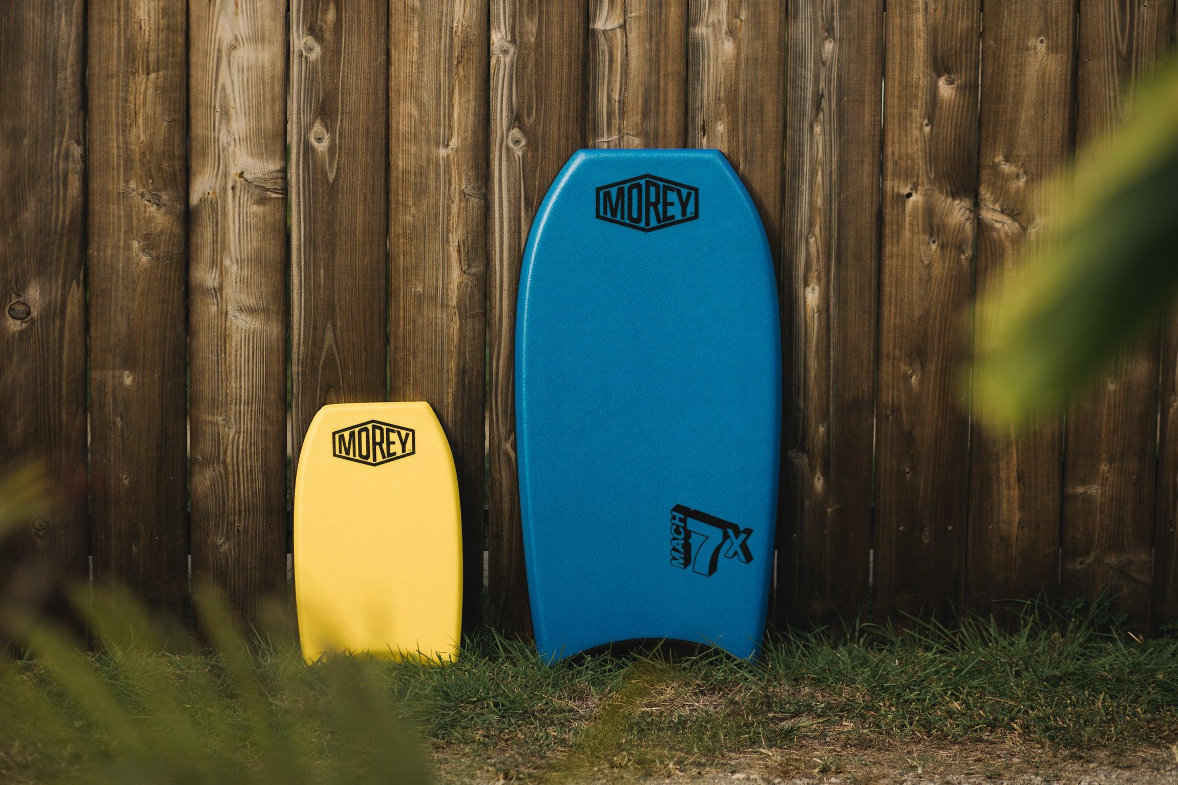 Yellow and blue bodyboards with Morey logos standing against wooden fence outdoors