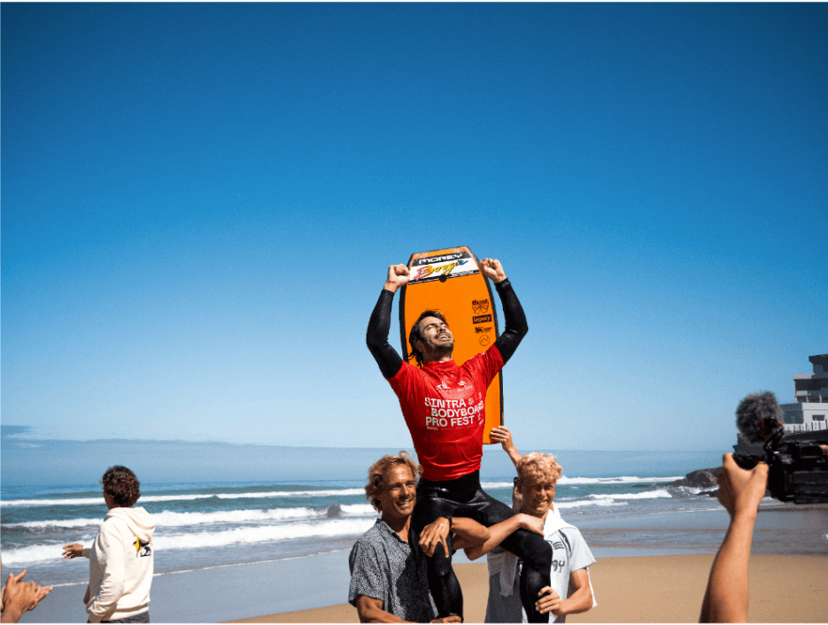 Winner holding orange bodyboard above head on beach during Sintra Bodyboard Pro Fest with ocean waves and cameraman