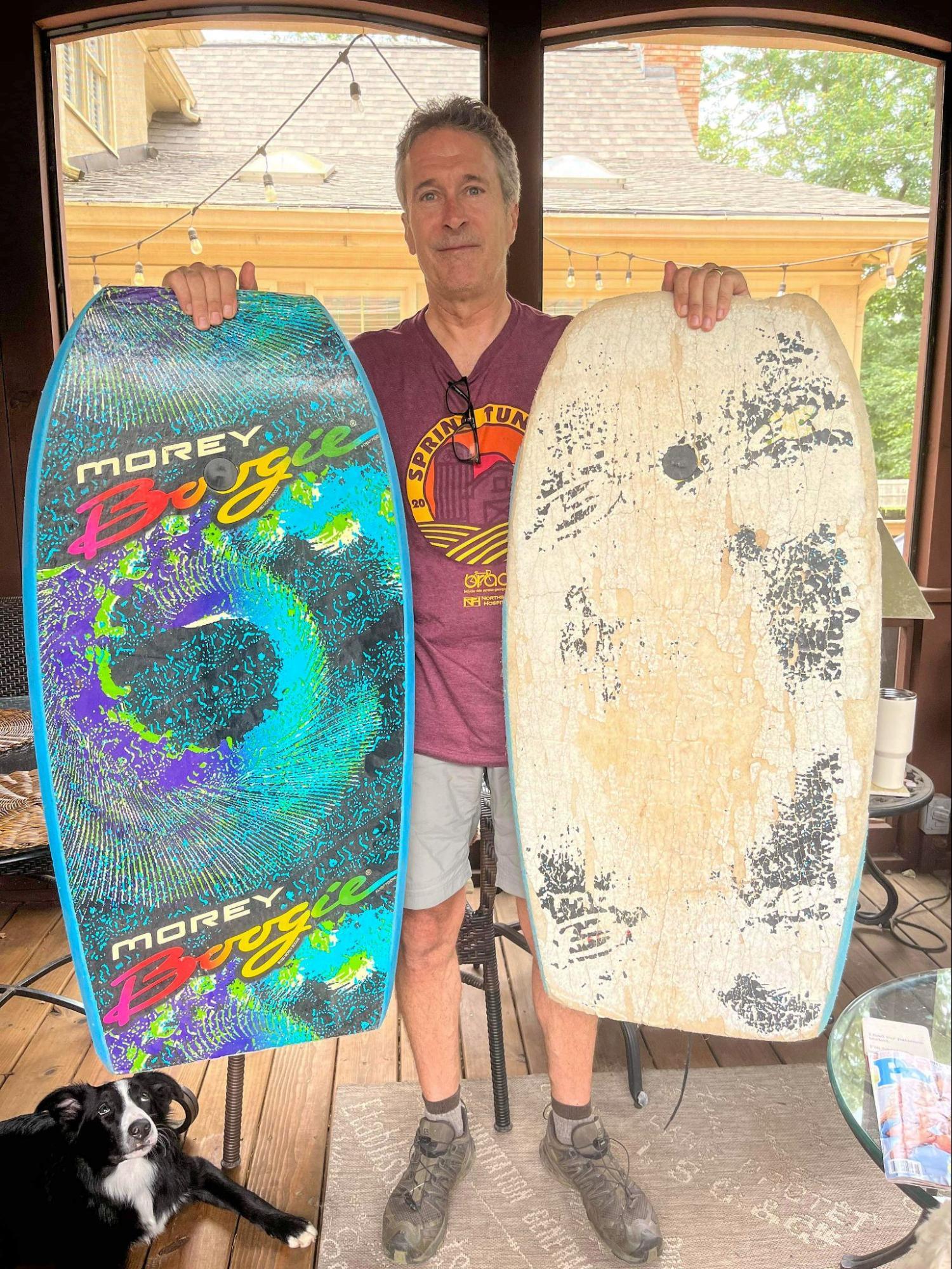 Man standing on wooden porch holding two Morey Boogie bodyboards, one vibrant and one worn, with black and white dog nearby