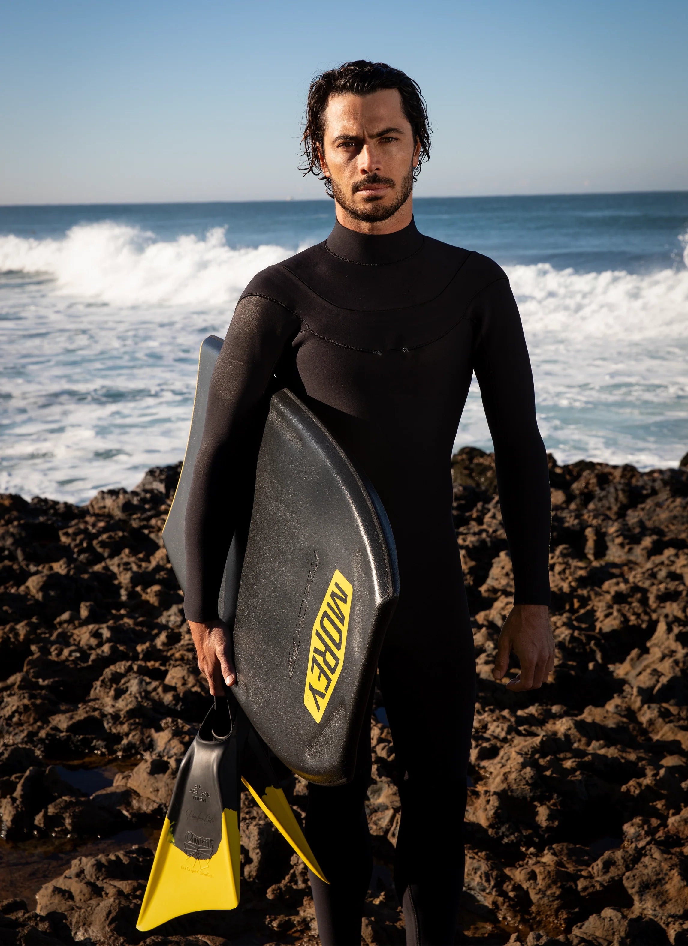 Man in wetsuit holding black bodyboard and yellow fins on rocky beach with ocean waves